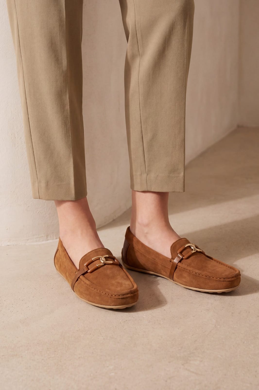 A person wearing tan suede loafers with gold-tone buckle details, paired with beige trousers, standing on a neutral concrete floor.