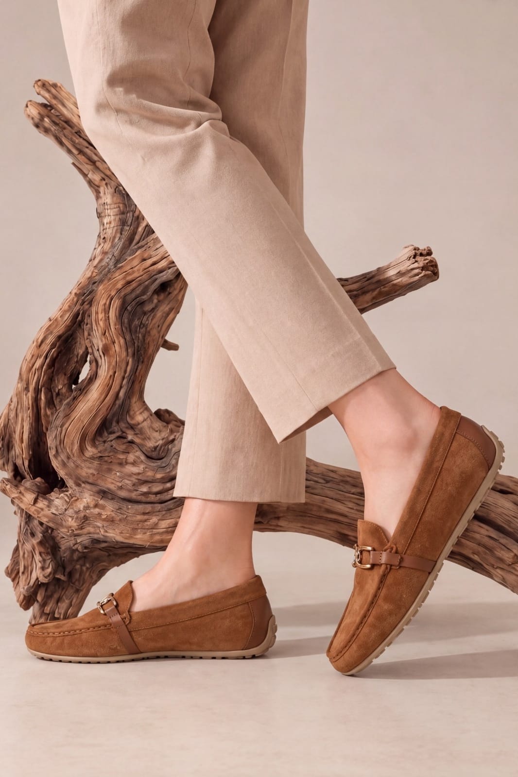 A person wearing tan suede moccasin loafers with a small gold-tone buckle detail, paired with beige trousers, seated on a piece of driftwood against a neutral beige background.