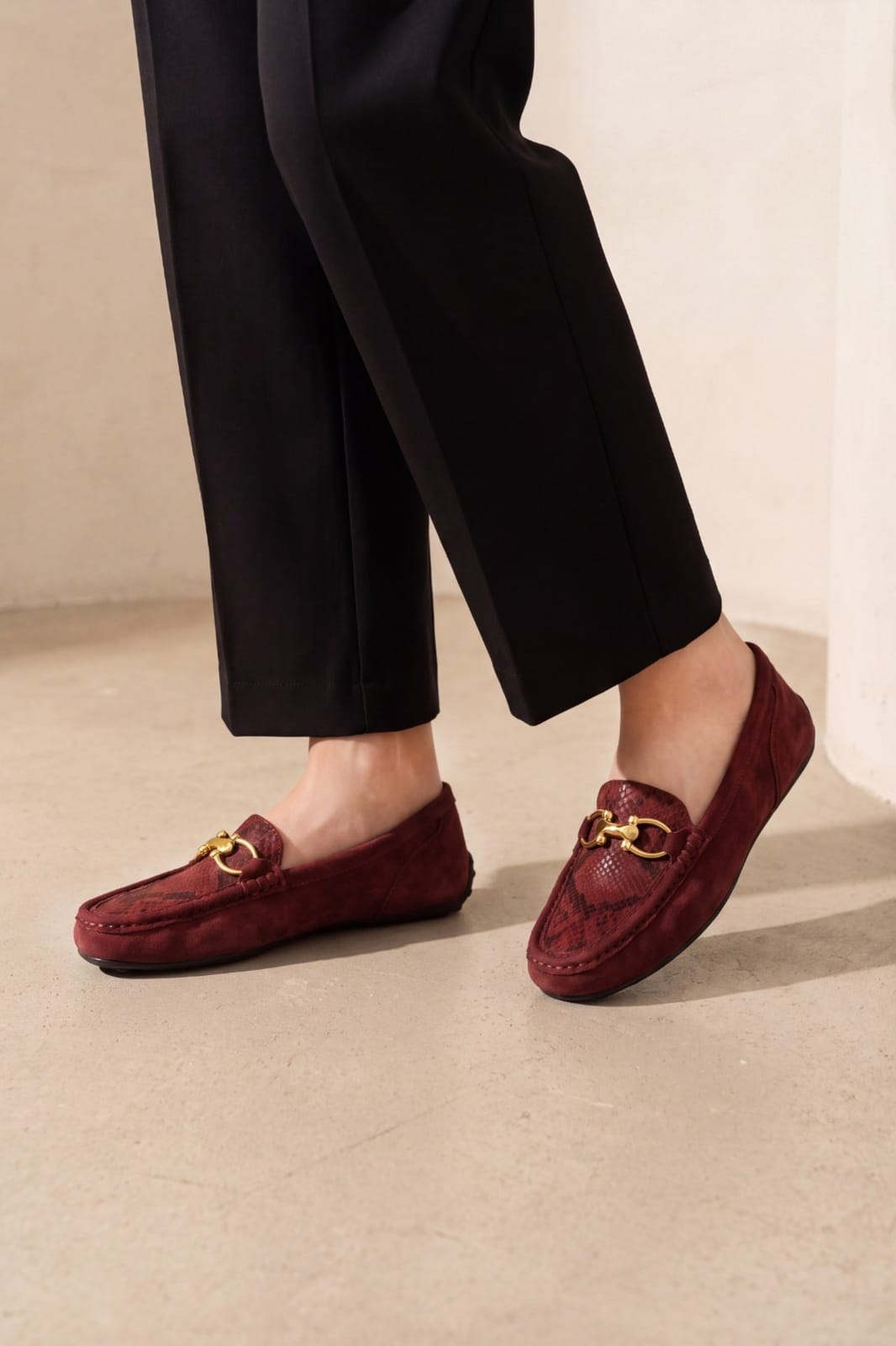 A person wearing burgundy snakeskin-patterned loafers with gold-tone horsebit details, paired with black trousers, standing on a light beige concrete floor.