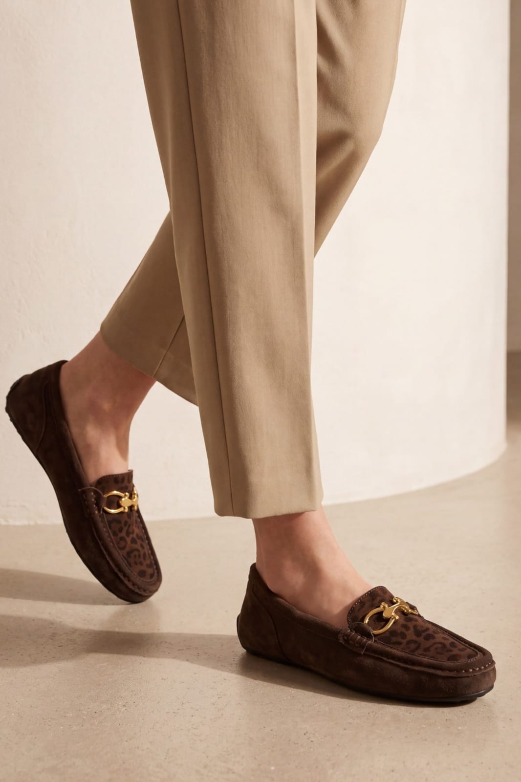 A person wearing brown suede and leopard-print loafers with gold horsebit details, paired with beige trousers, in a minimalist indoor setting.
