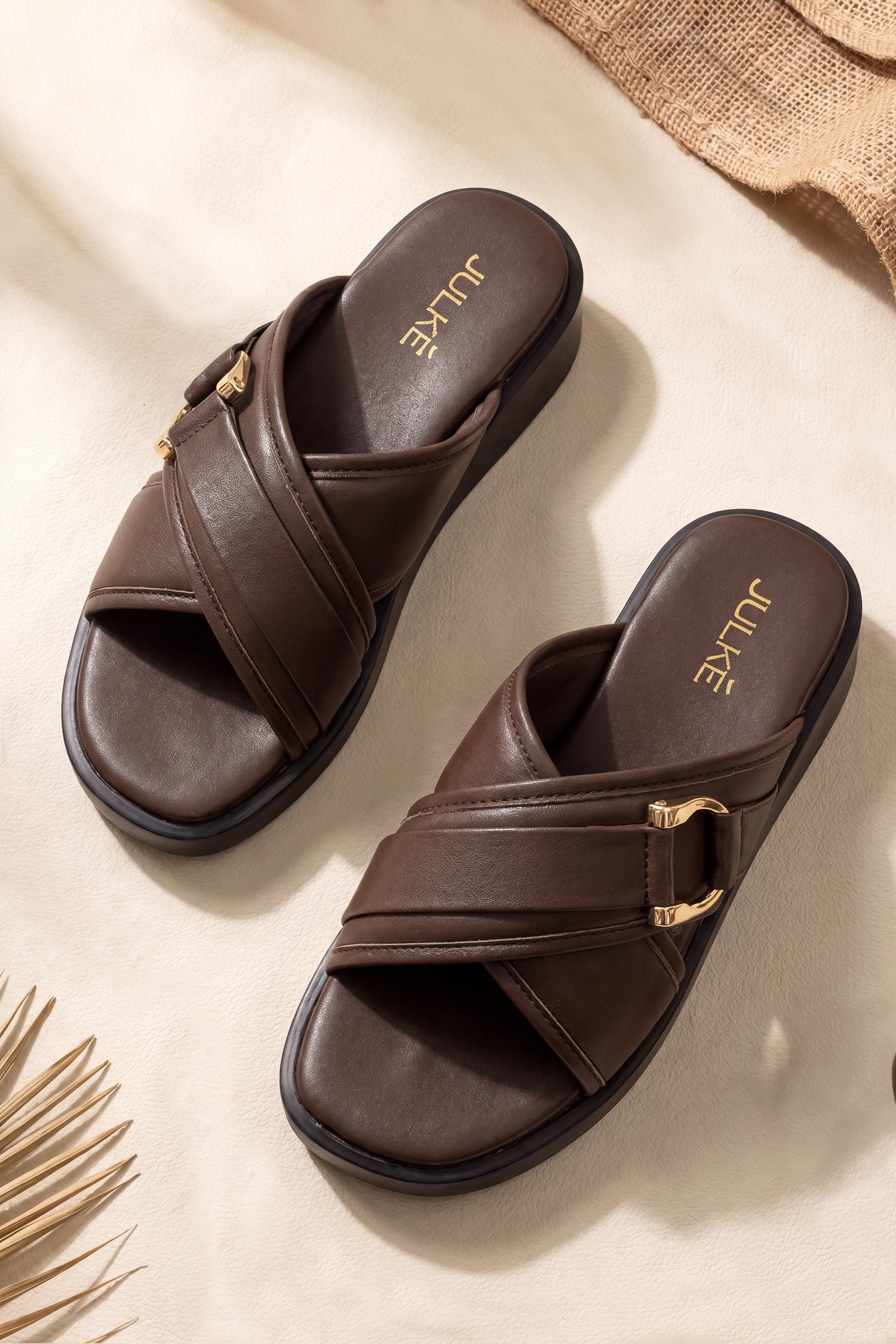 A pair of dark brown leather slide sandals with gold-tone buckle details, displayed on a textured beige surface with decorative straw elements.