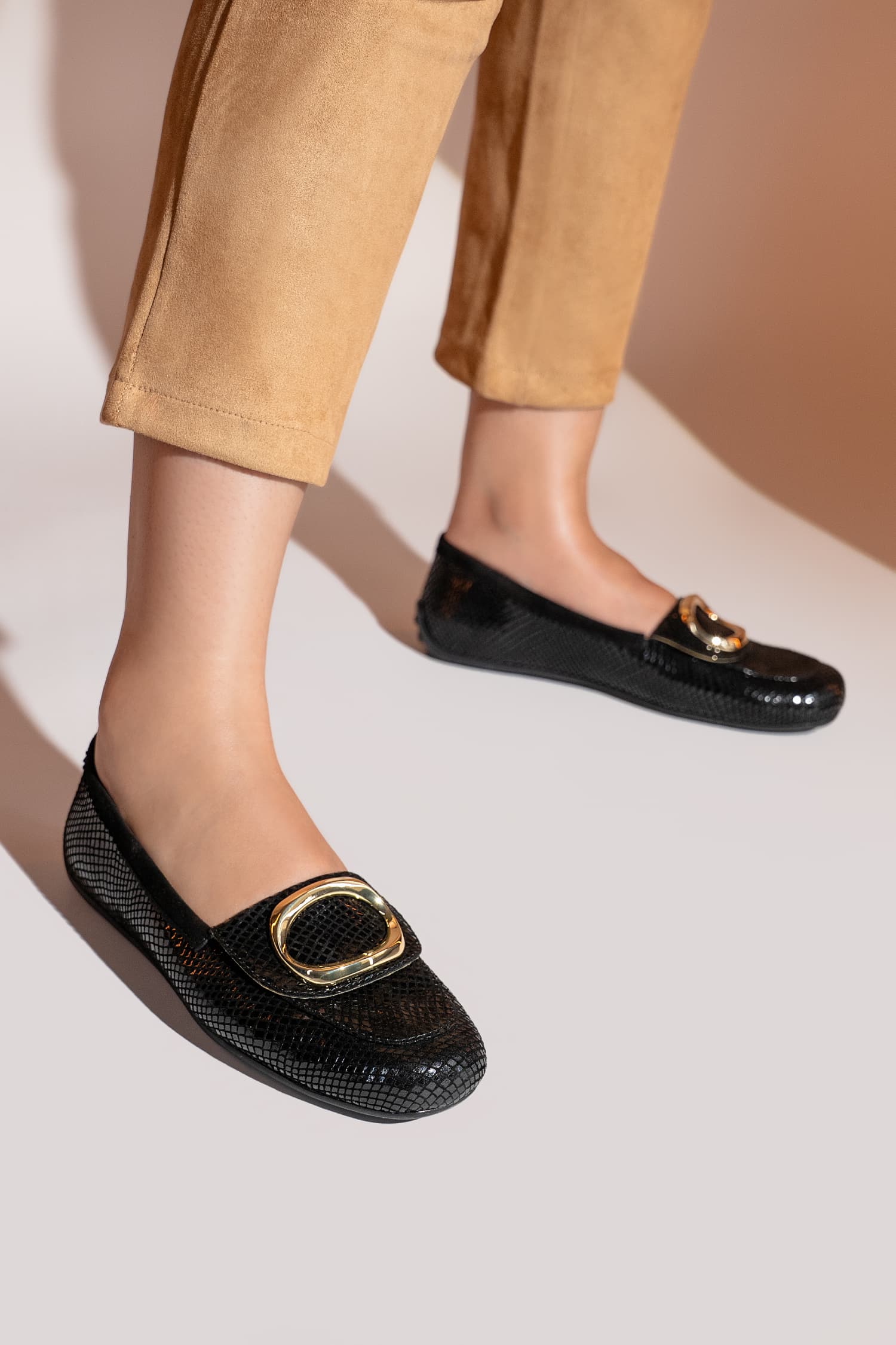 Black loafers with gold buckles worn by a person in beige pants on a light background