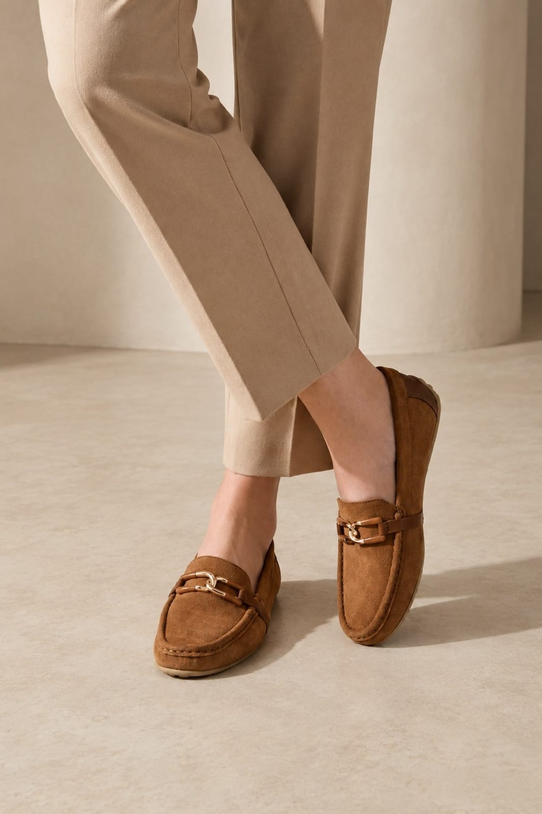 A person wearing tan suede loafers with gold-tone buckle details, paired with beige trousers, standing in a minimalist beige interior setting.