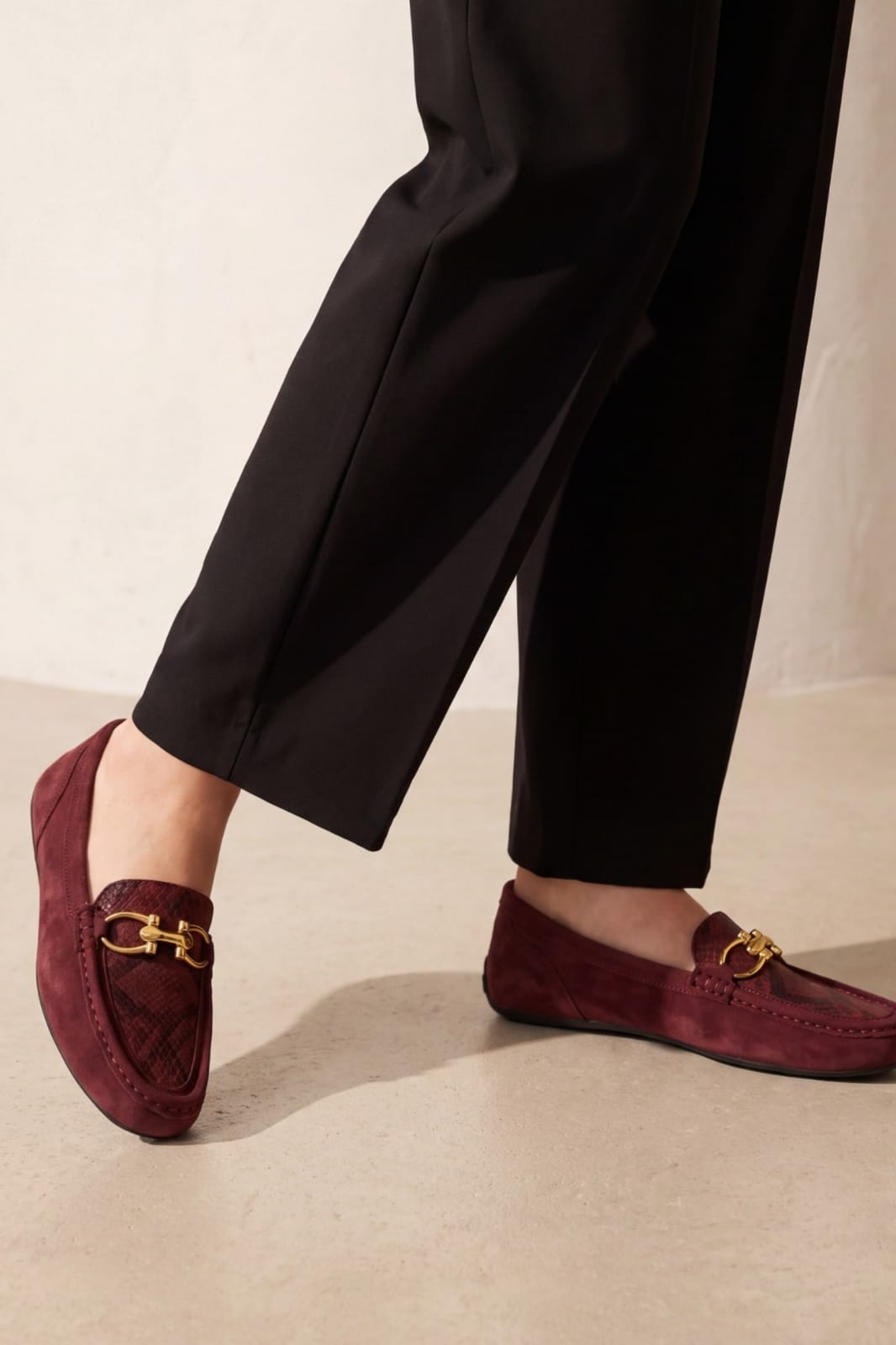 A close-up of a person's legs wearing black trousers and burgundy suede loafers with gold horsebit hardware, standing on a light beige floor against a neutral beige wall.