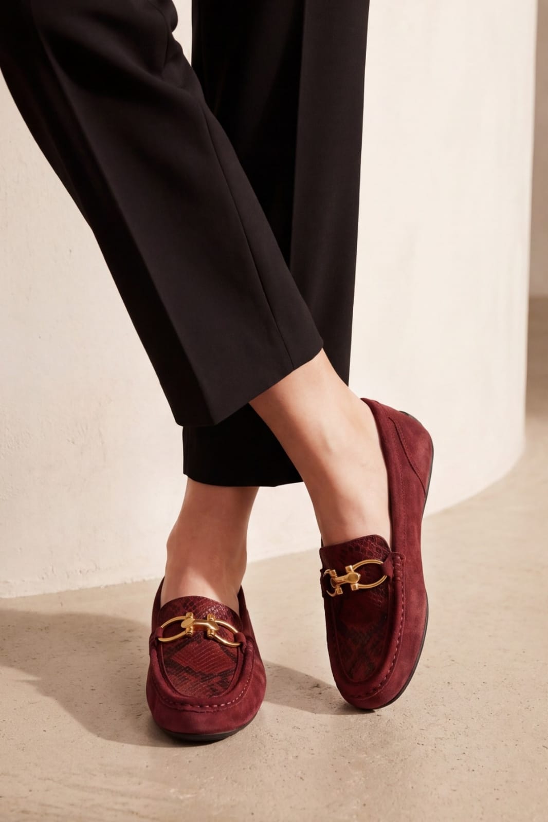 A person wearing burgundy suede loafers with gold horsebit hardware and black trousers, standing on a light concrete surface against a neutral wall.