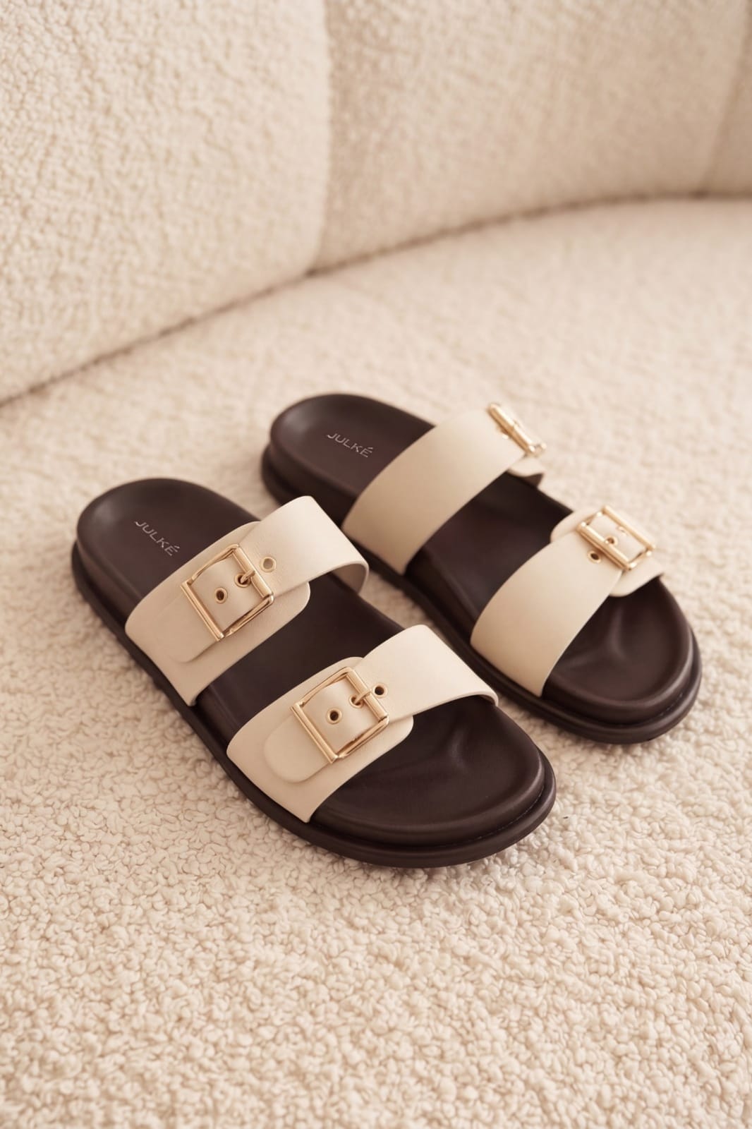 JULKÉ Luma comfort slide in ivory from Cloud Walk collection with gold buckle straps and brown footbed - flat lay on bouclé sofa