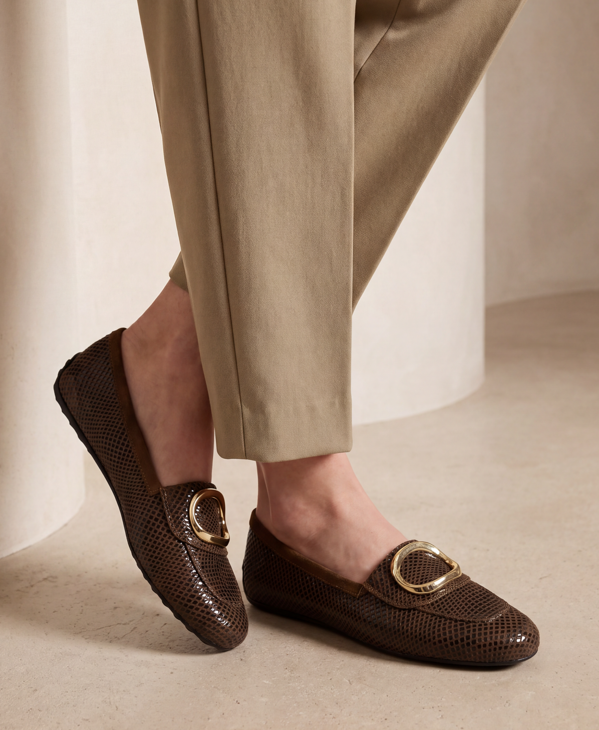 A person wearing brown textured slip-on loafers with gold-tone oval hardware and beige trousers, shown from the calves down in a softly lit indoor setting.